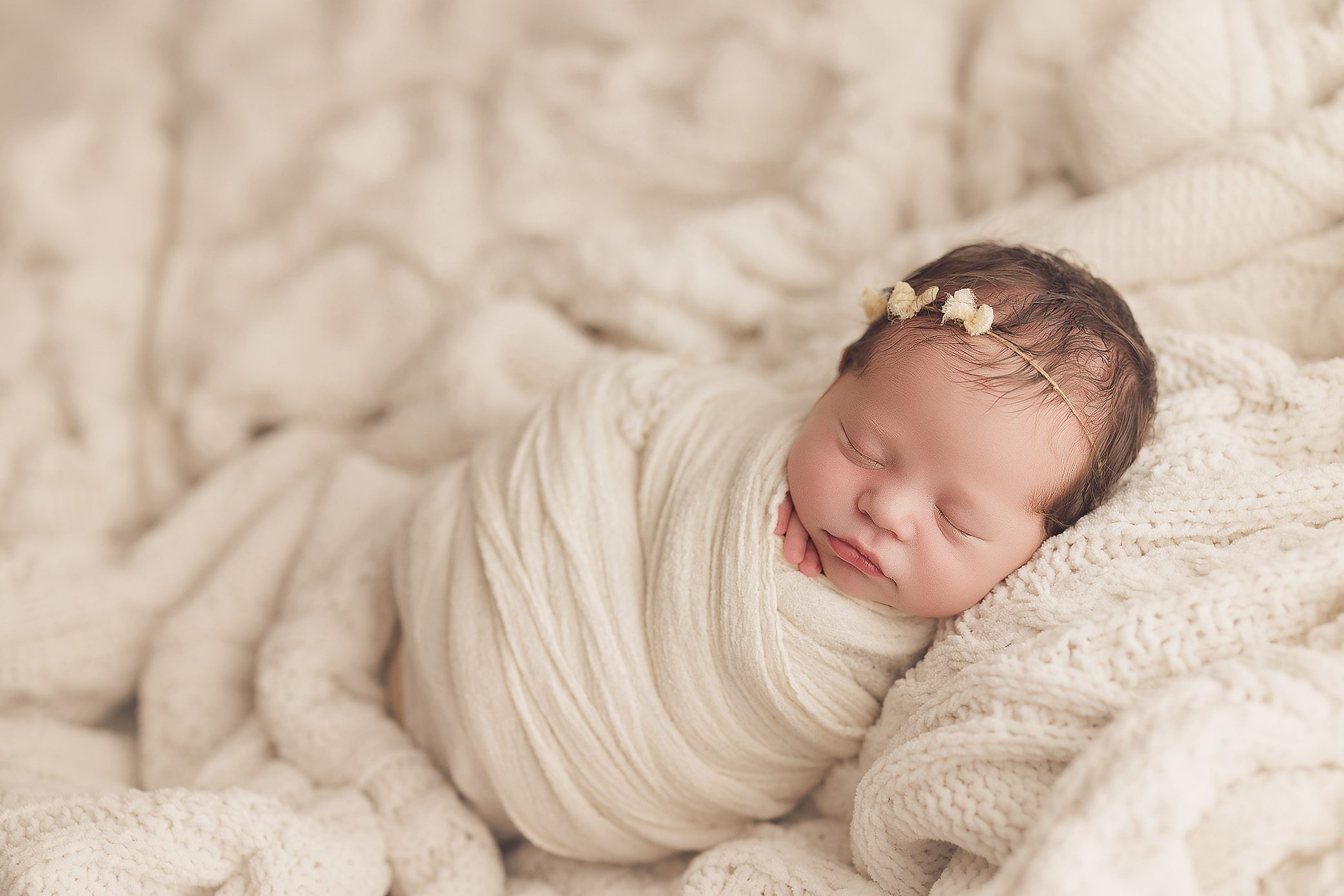 Newborn baby girl swaddled in a cream wrap, peacefully sleeping on a textured knit blanket with a delicate floral headband.