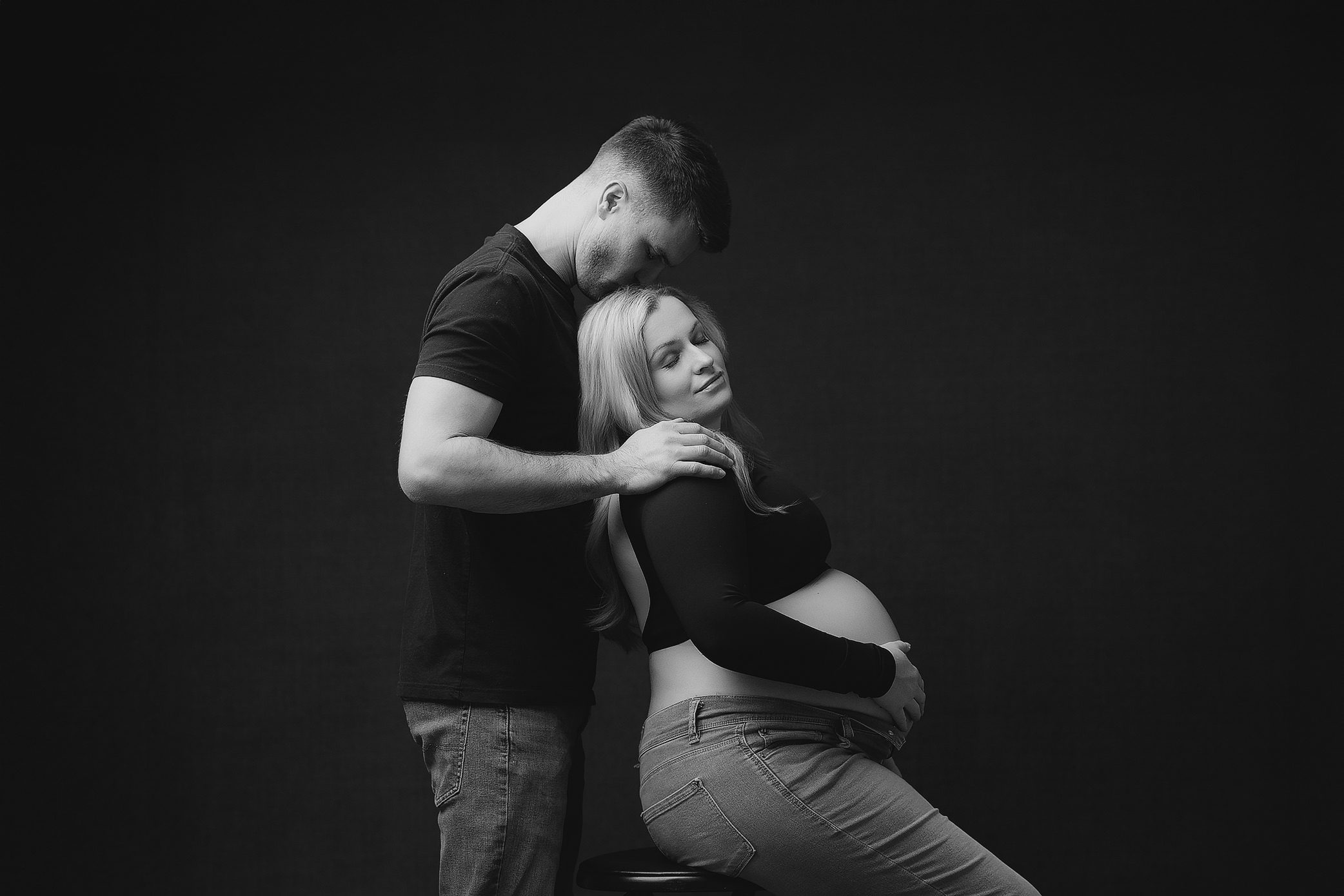 Black and white portrait of an expecting couple. Woman is sitting on a stool with her pregnant belly bare, and the man is kissing the top of her head with his hands on her shoulders.