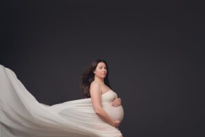 Studio maternity portrait of a pregnant woman in a cream gown with flowing fabric against a dark backdrop