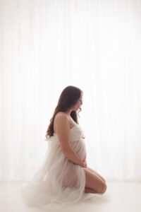 Pregnant woman seated in profile wearing a sheer white maternity gown in a softly lit studio