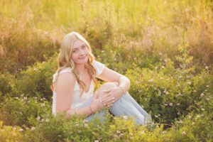 Close outdoor portrait of a smiling senior seated in greenery with soft golden backlighting.