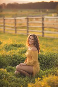 Smiling student seated in tall grass beside split-rail fencing in a mustard floral top and jeans.