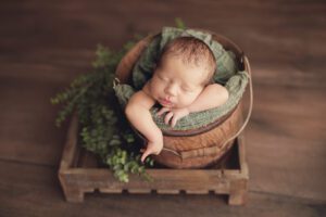 Baby boy posed in a bucket on a wood floor with greenery at a Rockville newborn photography studio